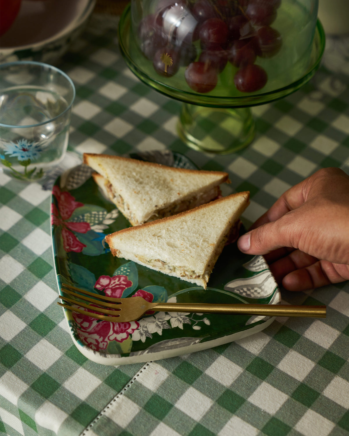 Hand holding a Buransh floral platter with sandwiches and a gold fork.