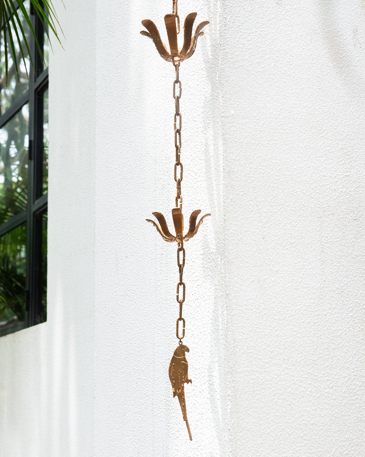 Madagascar Rain Chain with two flower-shaped cups and a parrot pendant against a white wall.