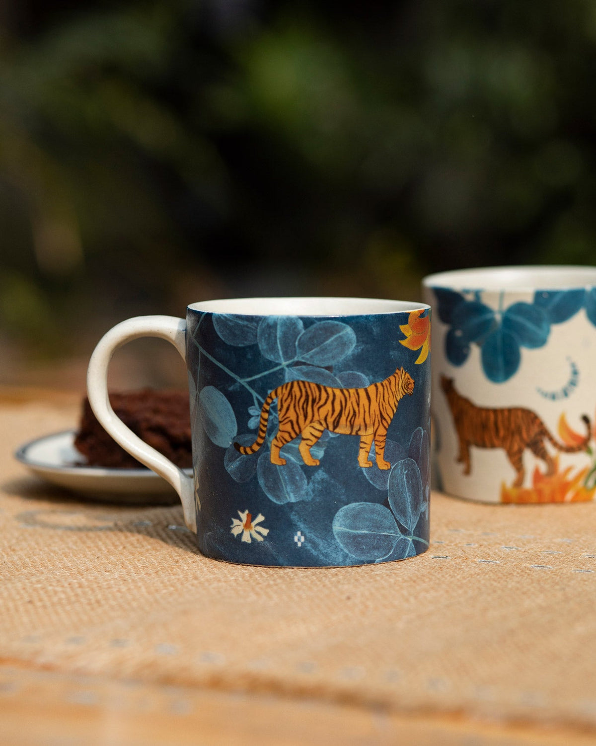 Navy Nadi mug with an orange tiger among blue leaves, cream mug behind.