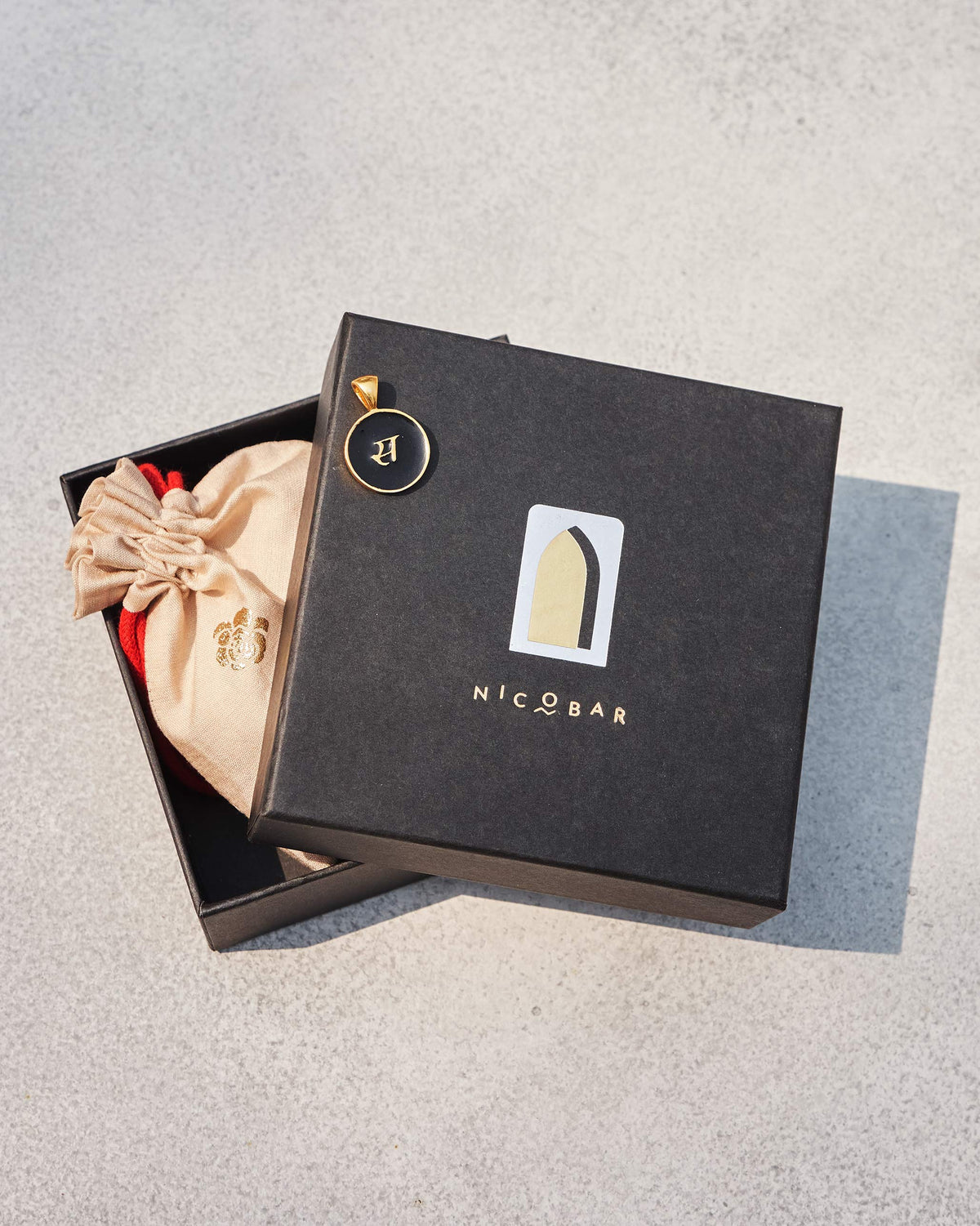 Black enamel charm with gold Devanagari स on a black Nicobar gift box.