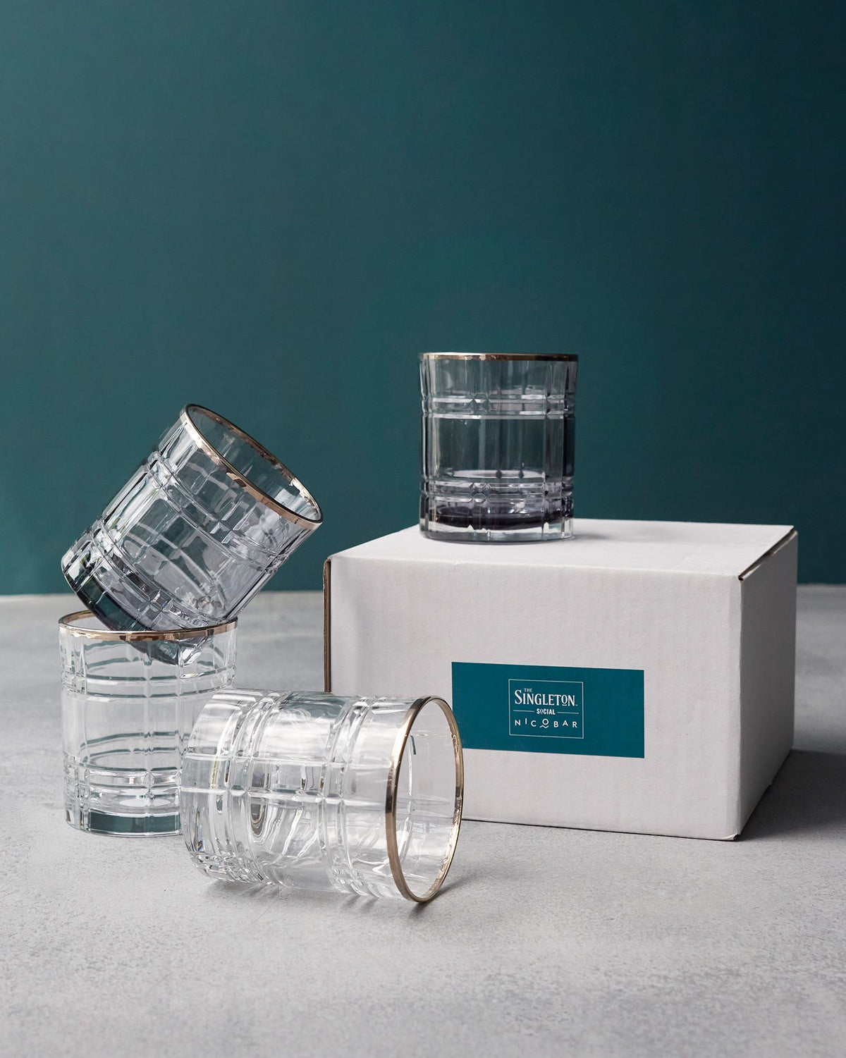 Four gold-rimmed Celeste whiskey glasses with a grid pattern beside their gift box.