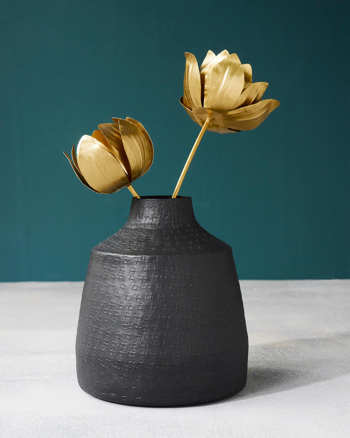 Dhaara Vase in black with a textured cross pattern, holding two gold flowers.
