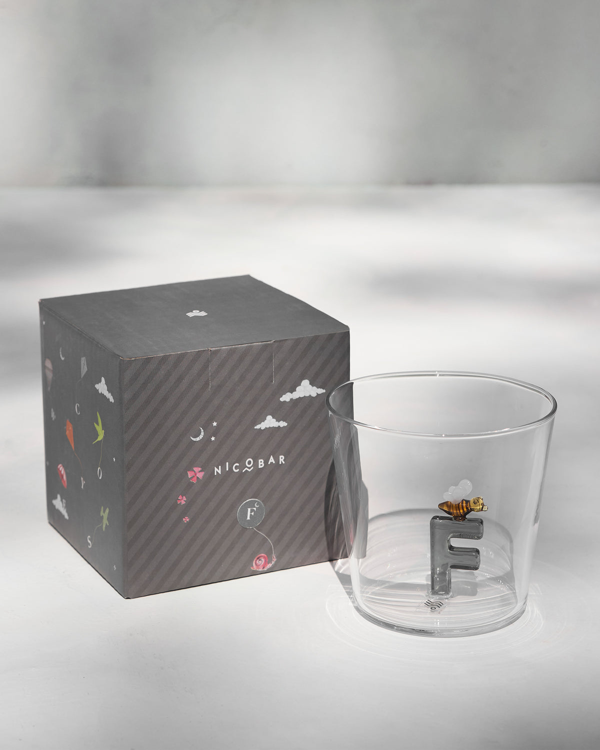 Clear Alphabet glass with a 3D letter F beside a charcoal Nicobar gift box.