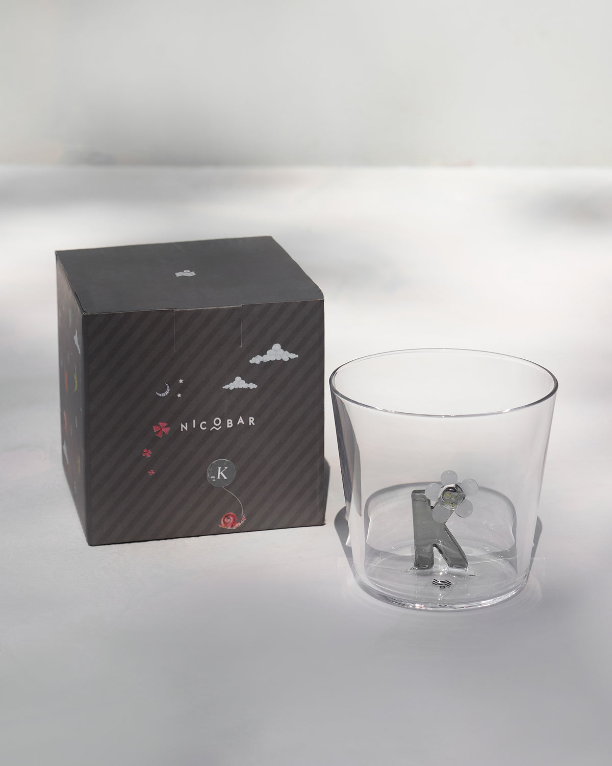 Alphabet glass with a silver letter K beside its charcoal Nicobar gift box.