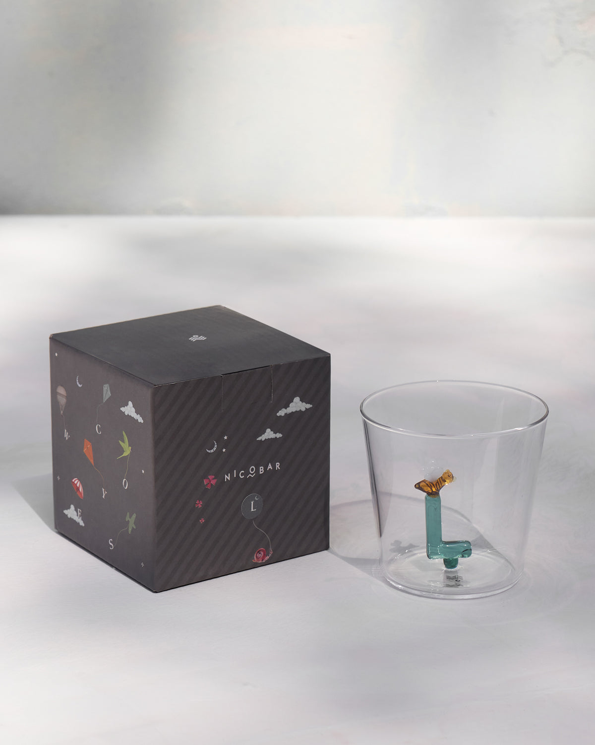 Alphabet glass with a teal letter L beside its charcoal Nicobar gift box.