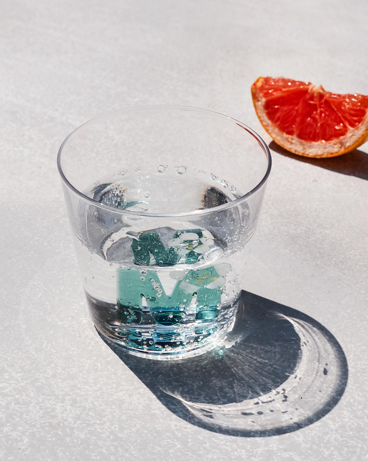 Overhead view of an Alphabet glass with a teal floral letter M in sparkling water.