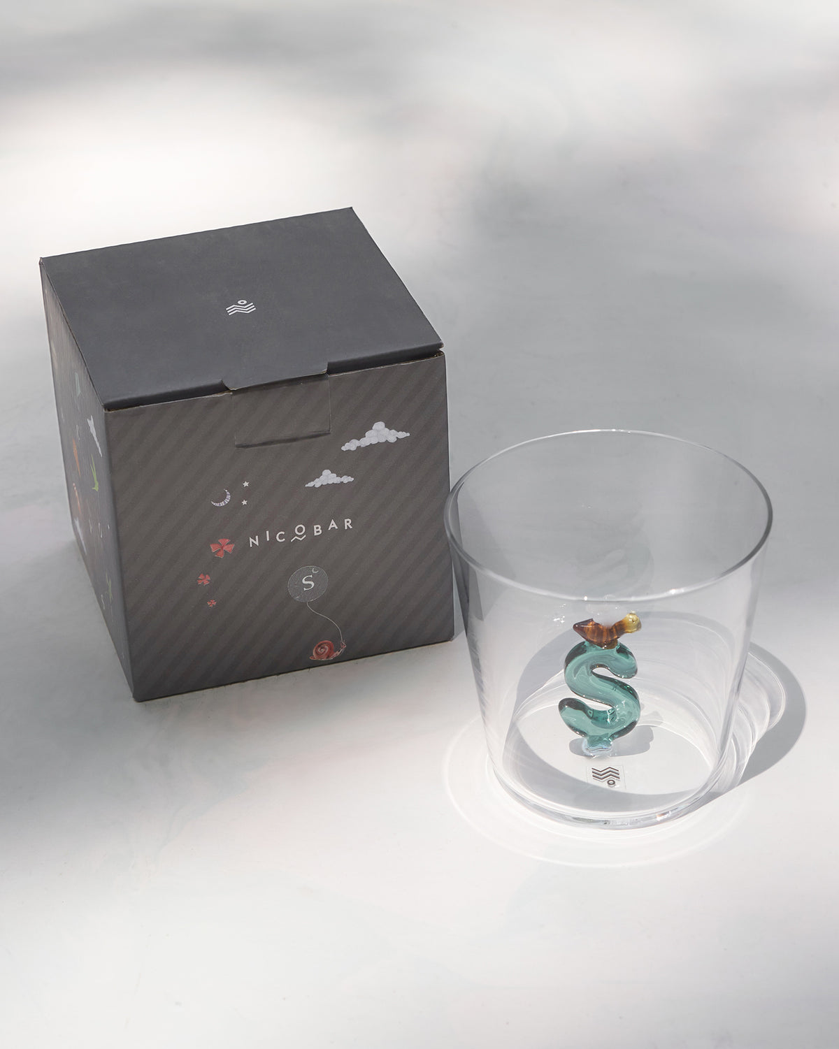 Alphabet glass with a teal letter S beside its charcoal Nicobar gift box.