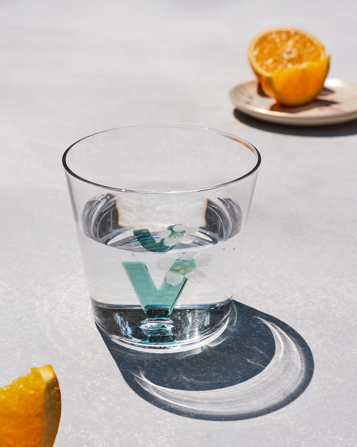 Clear Alphabet glass with a teal letter V and white flowers, half filled with water.