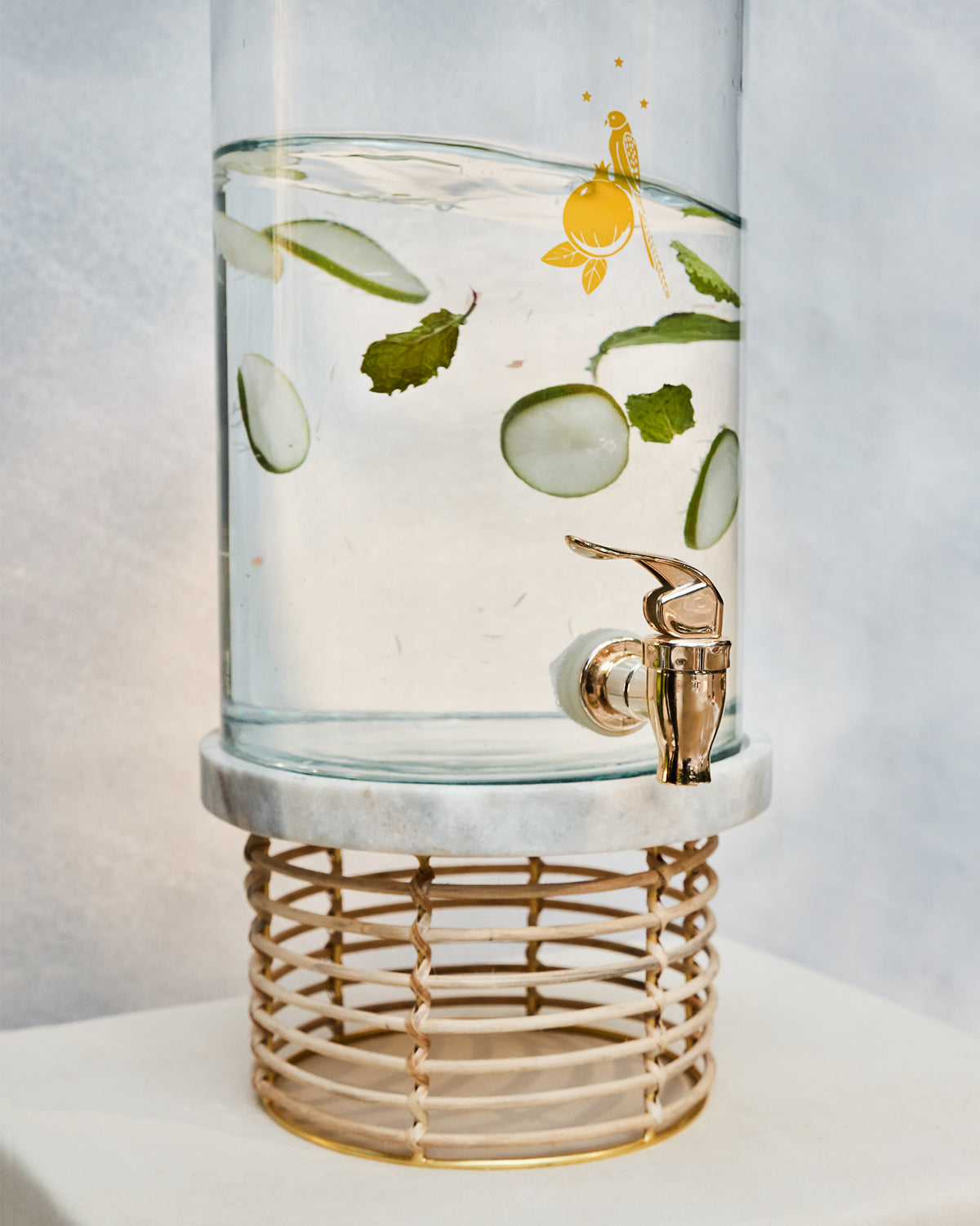 Mistle glass dispenser with gold tap on marble and rattan, filled with cucumber water.