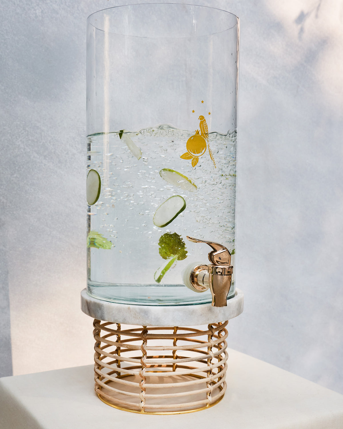 Mistle dispenser filled with cucumber and mint water on a rattan stand.