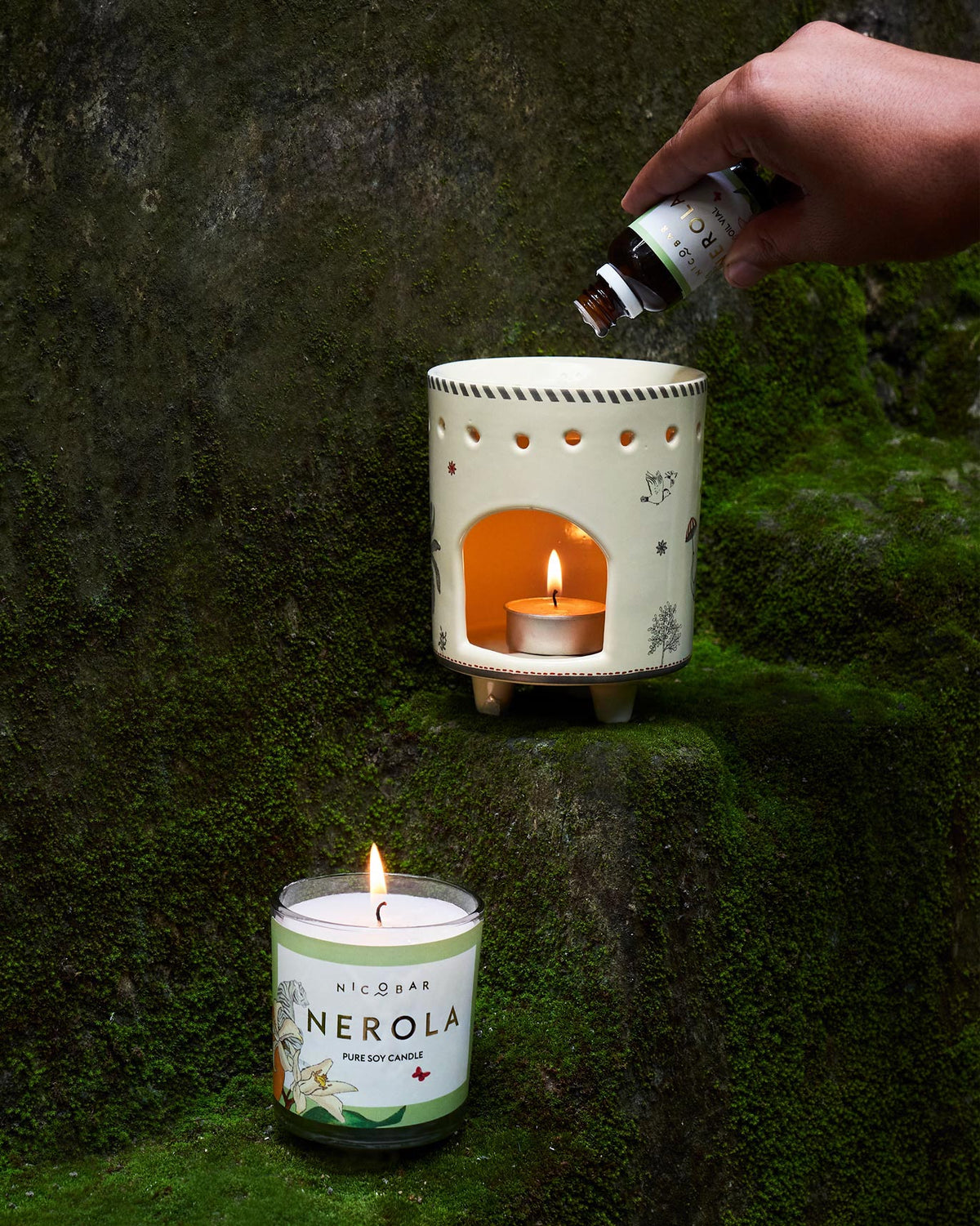 Person pouring oil from a vial into a ceramic burner beside a lit soy candle.