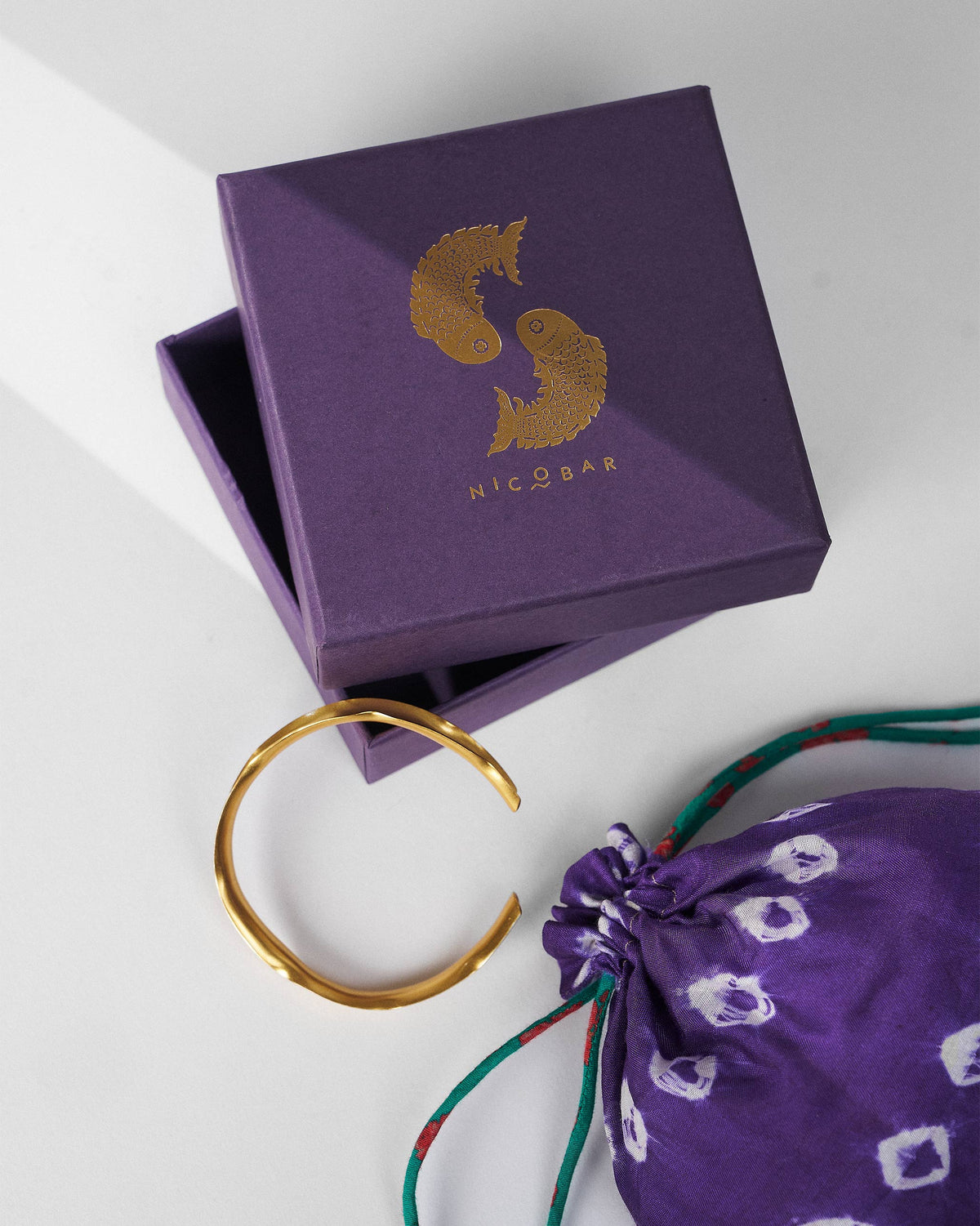 Thin gold Batur bangle beside a purple Nicobar gift box and pouch.