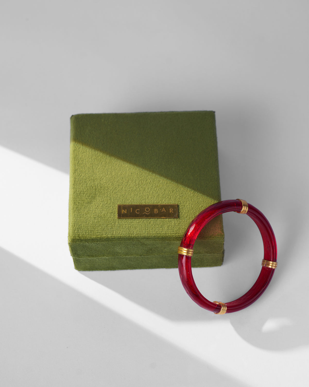 Red glass bangle with gold accents leaning on an olive velvet Nicobar box.