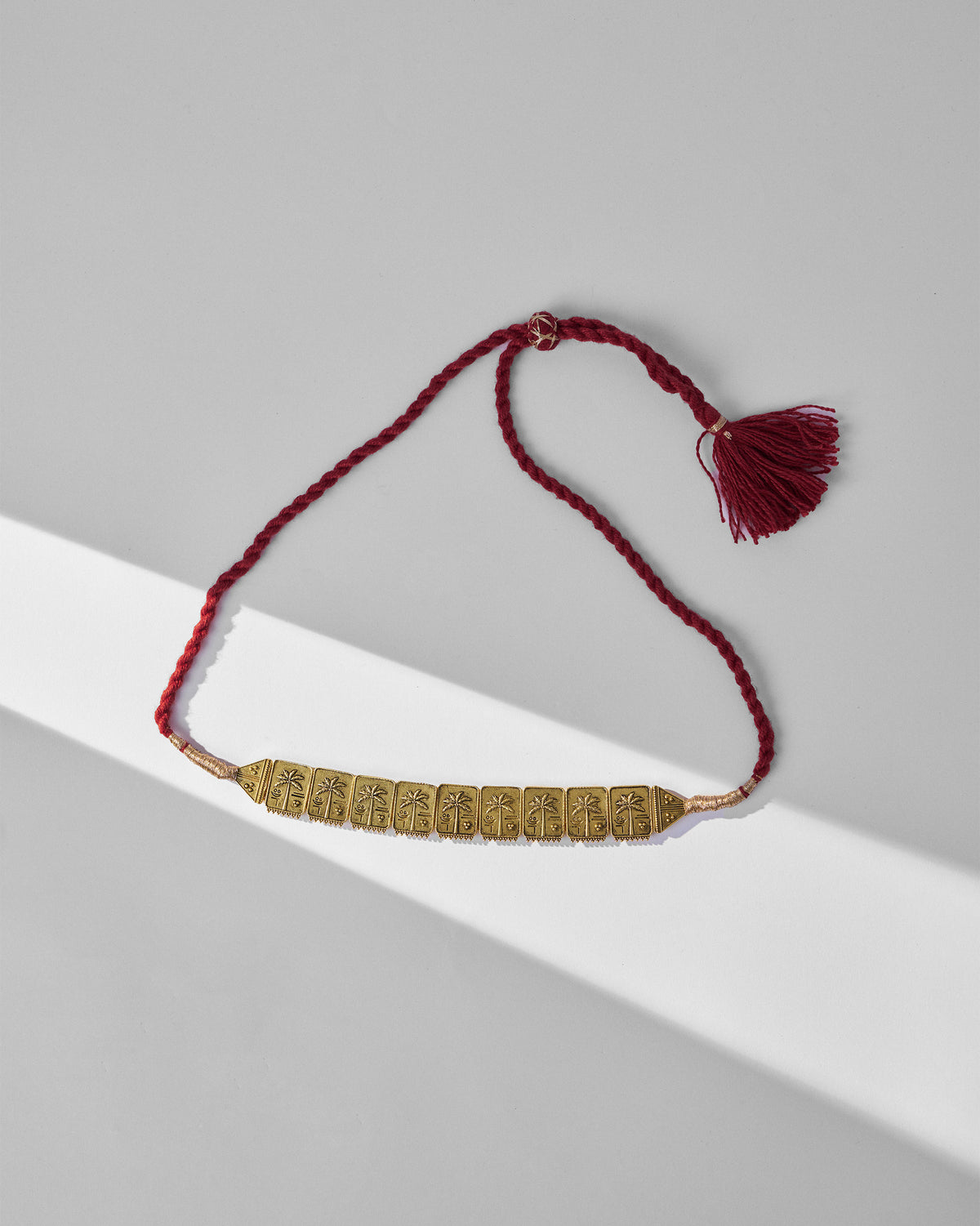 Antique gold coco palm necklace with maroon cord and tassel laid flat.