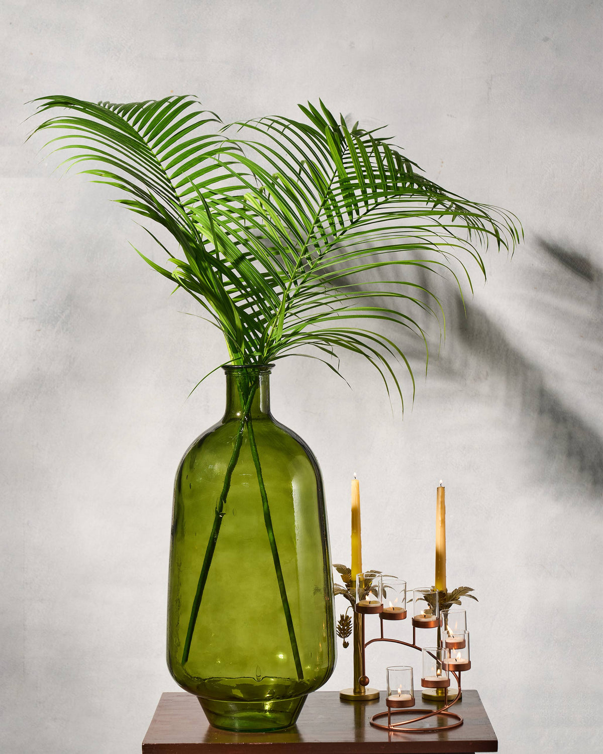Jade Glass Vase with palm fronds on a wood surface beside lit candles.