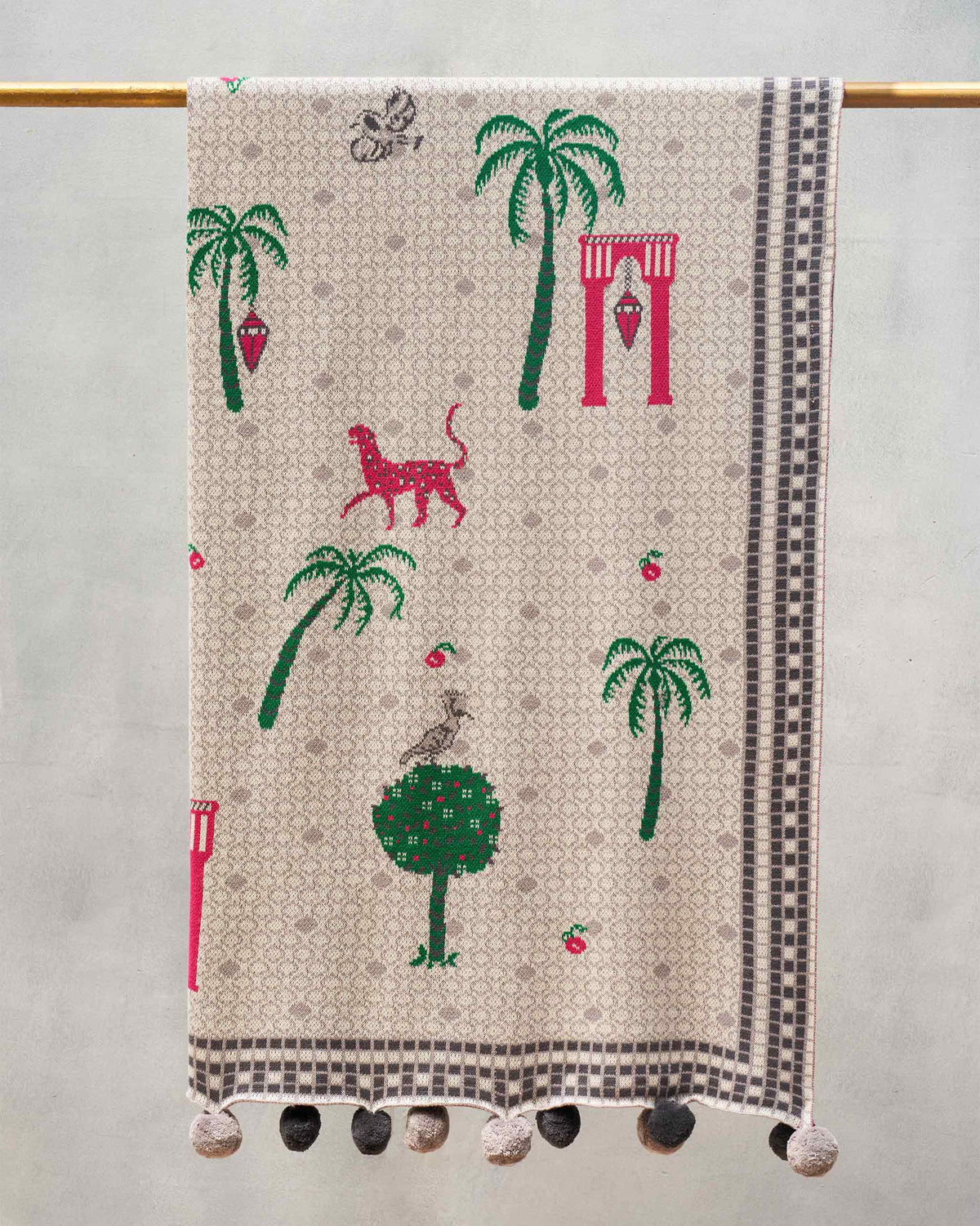 Mirage throw hanging with palm trees, a spotted cat and pom-pom edges.