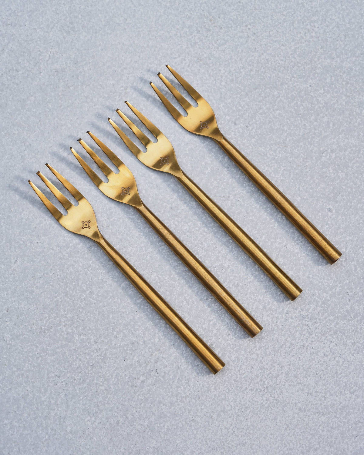 Four gold Sahara dessert forks arranged diagonally on a grey surface.