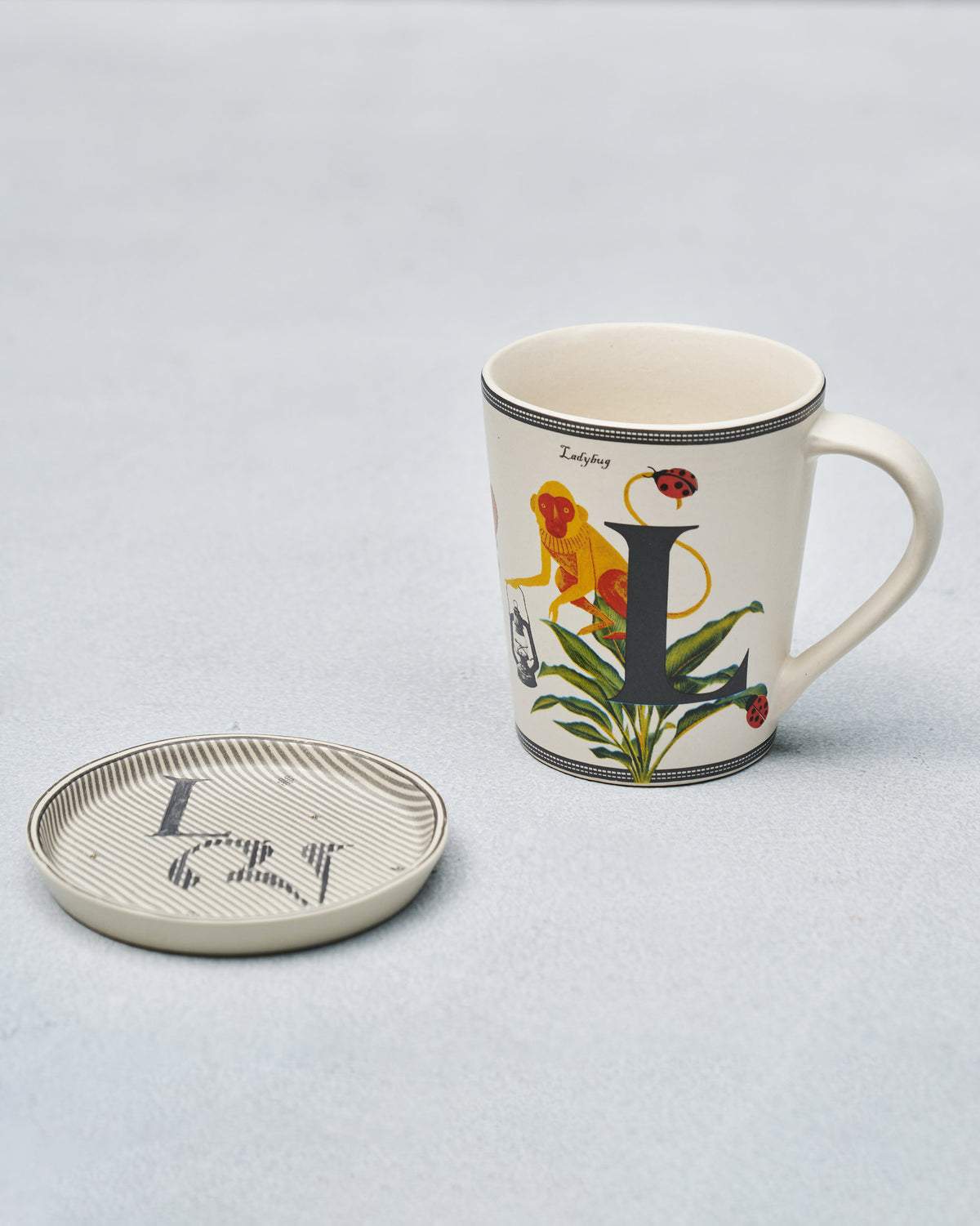 Cream L alphabet mug with monkey and ladybug beside matching coaster.