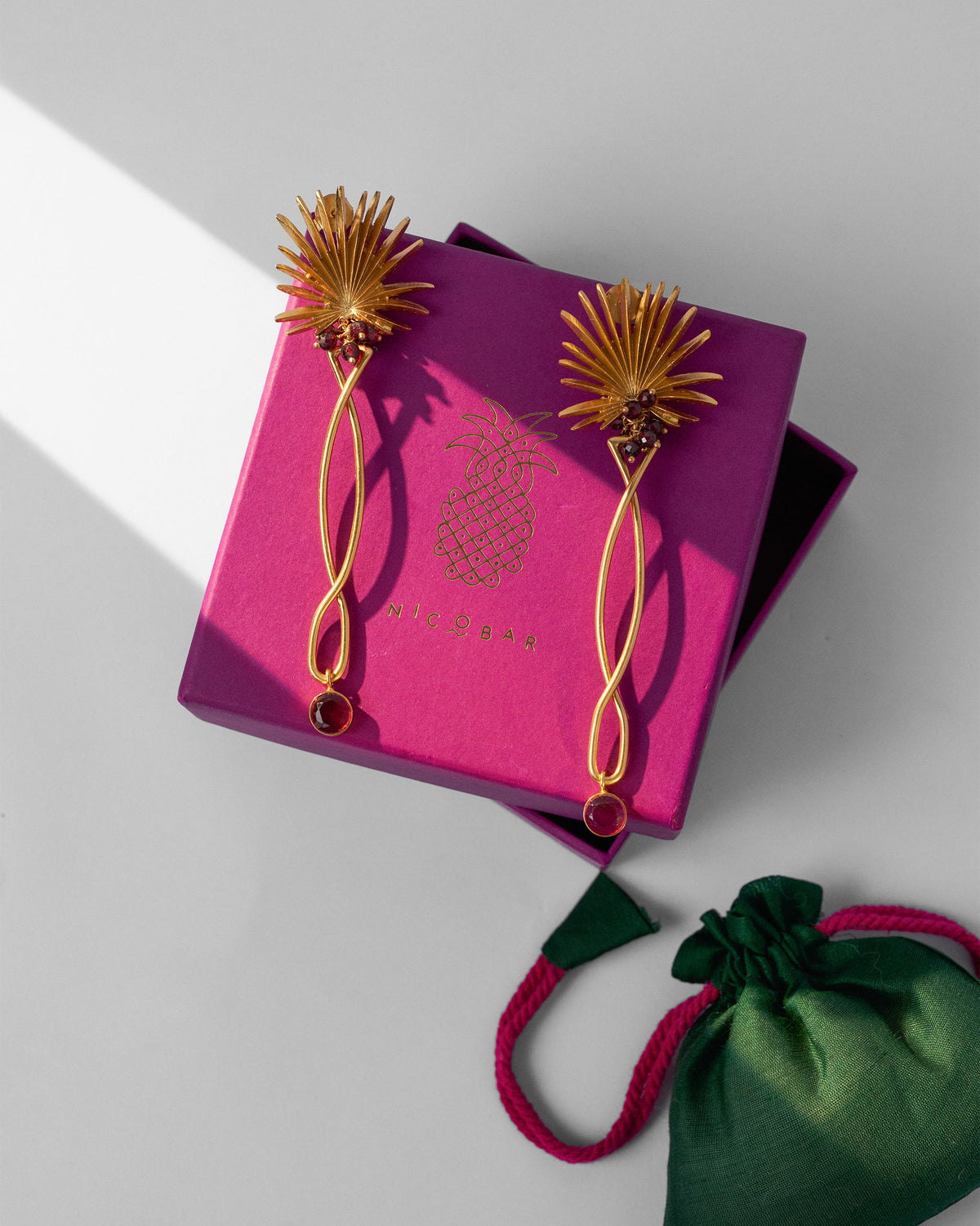 Pair of gold Palm Danglers on a pink gift box beside a green pouch.