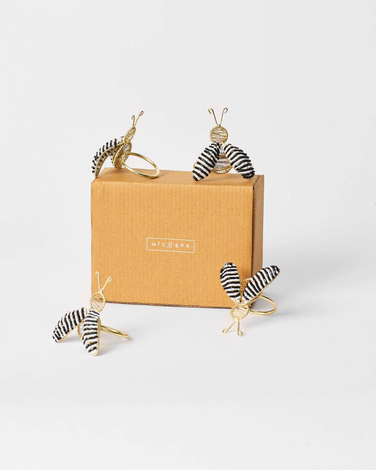 Four gold bee napkin rings with striped wings around a brown Nicobar box.