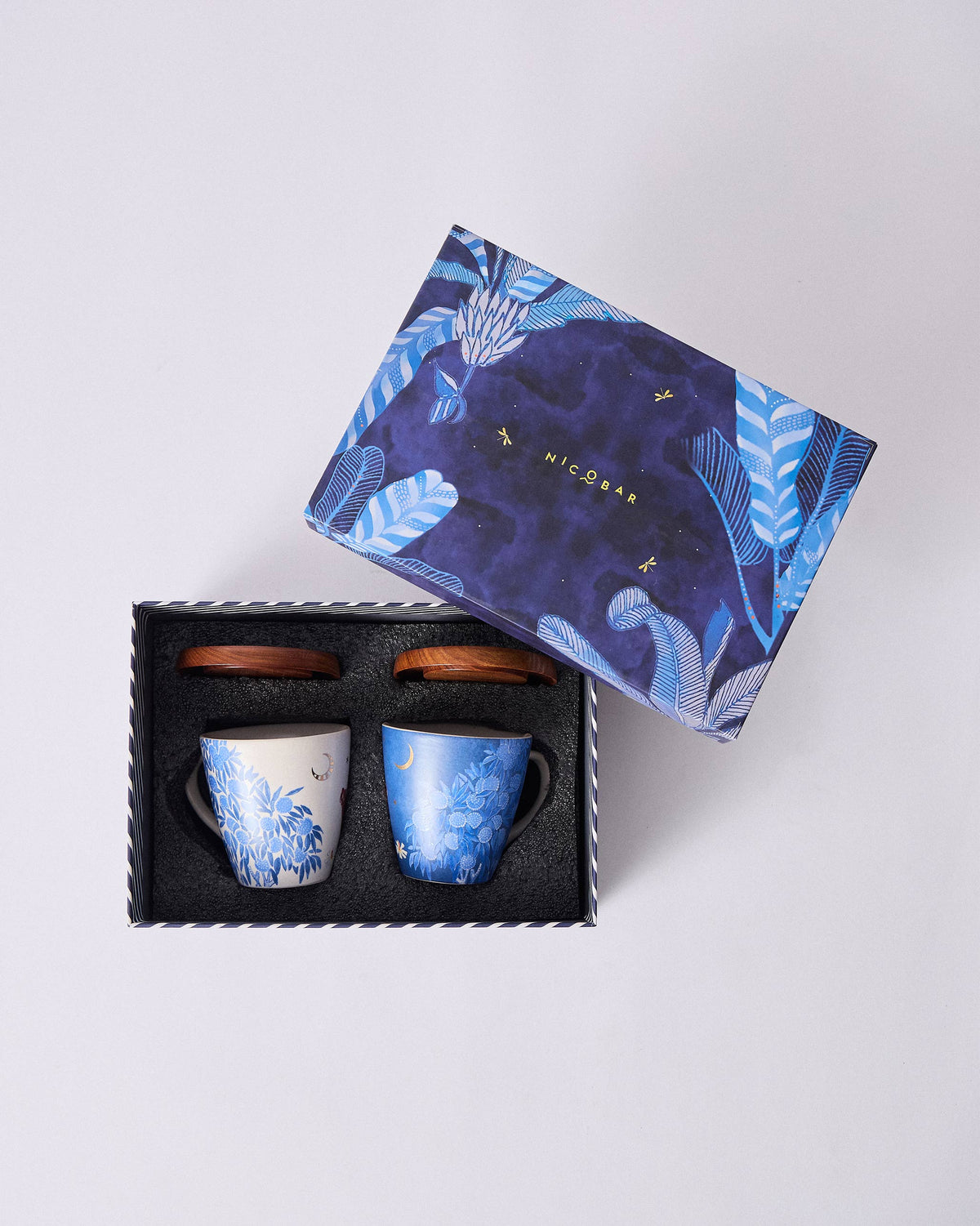 Open Nadi gift box with two botanical mugs and matching wooden lids in foam.