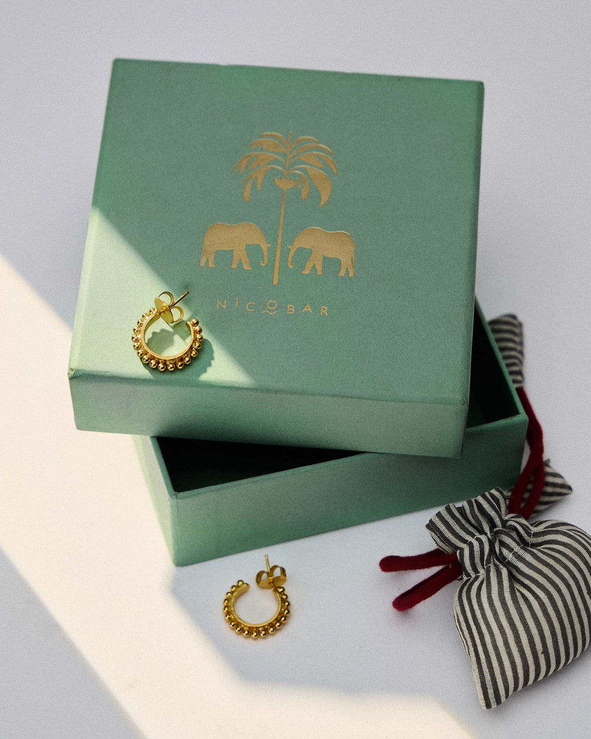 Pair of gold Rava Hoops Small on a green gift box beside a striped pouch.
