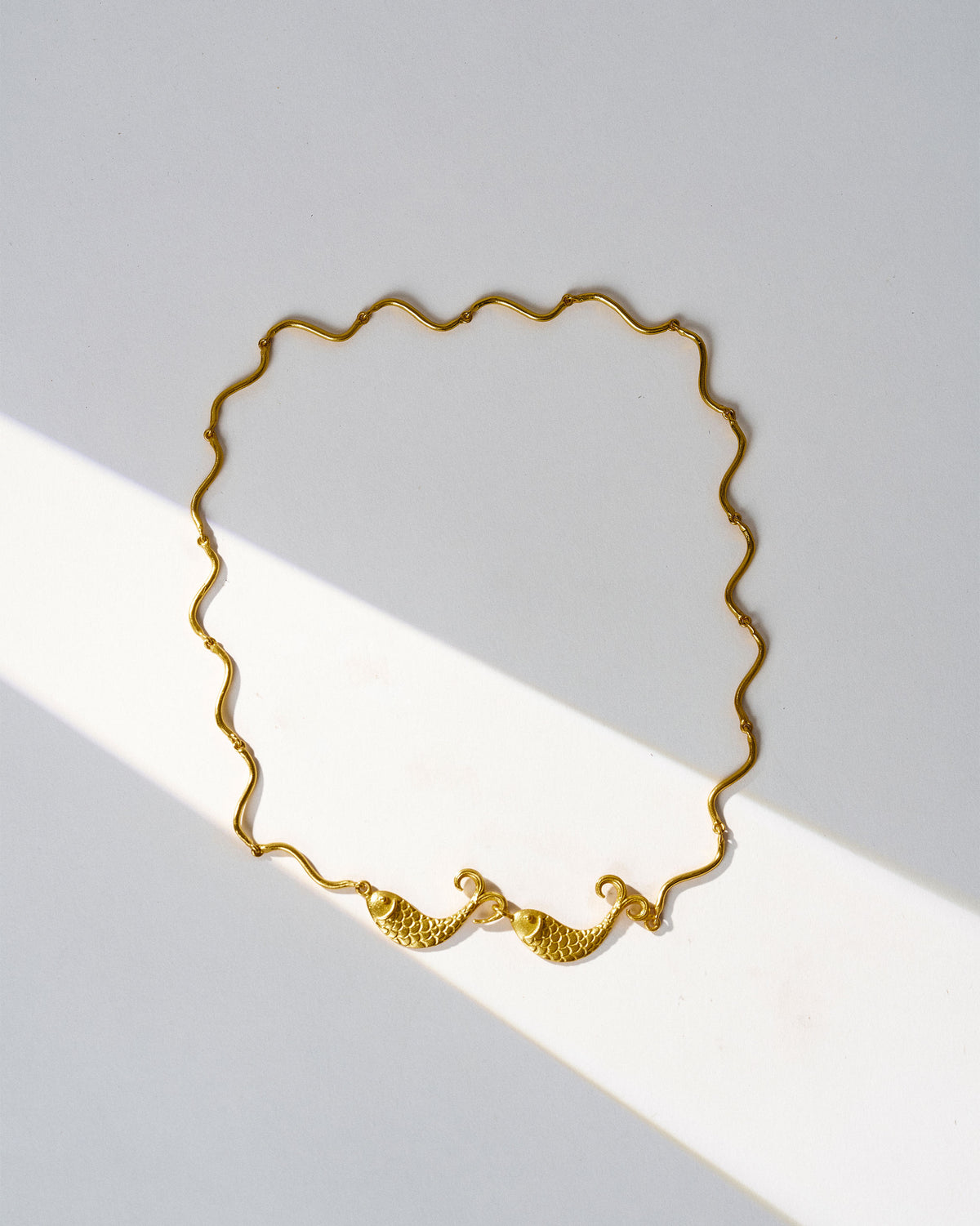 Gold Meen necklace with wavy chain and twin fish laid flat.
