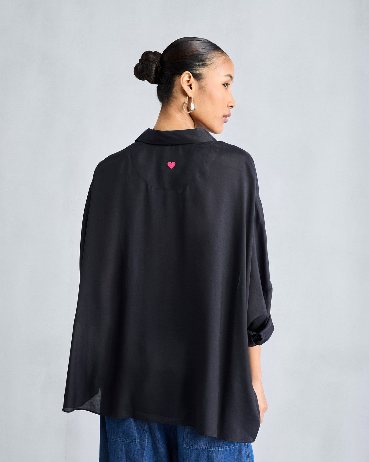 Back of a black boxy shirt with a pink embroidered heart below the collar.
