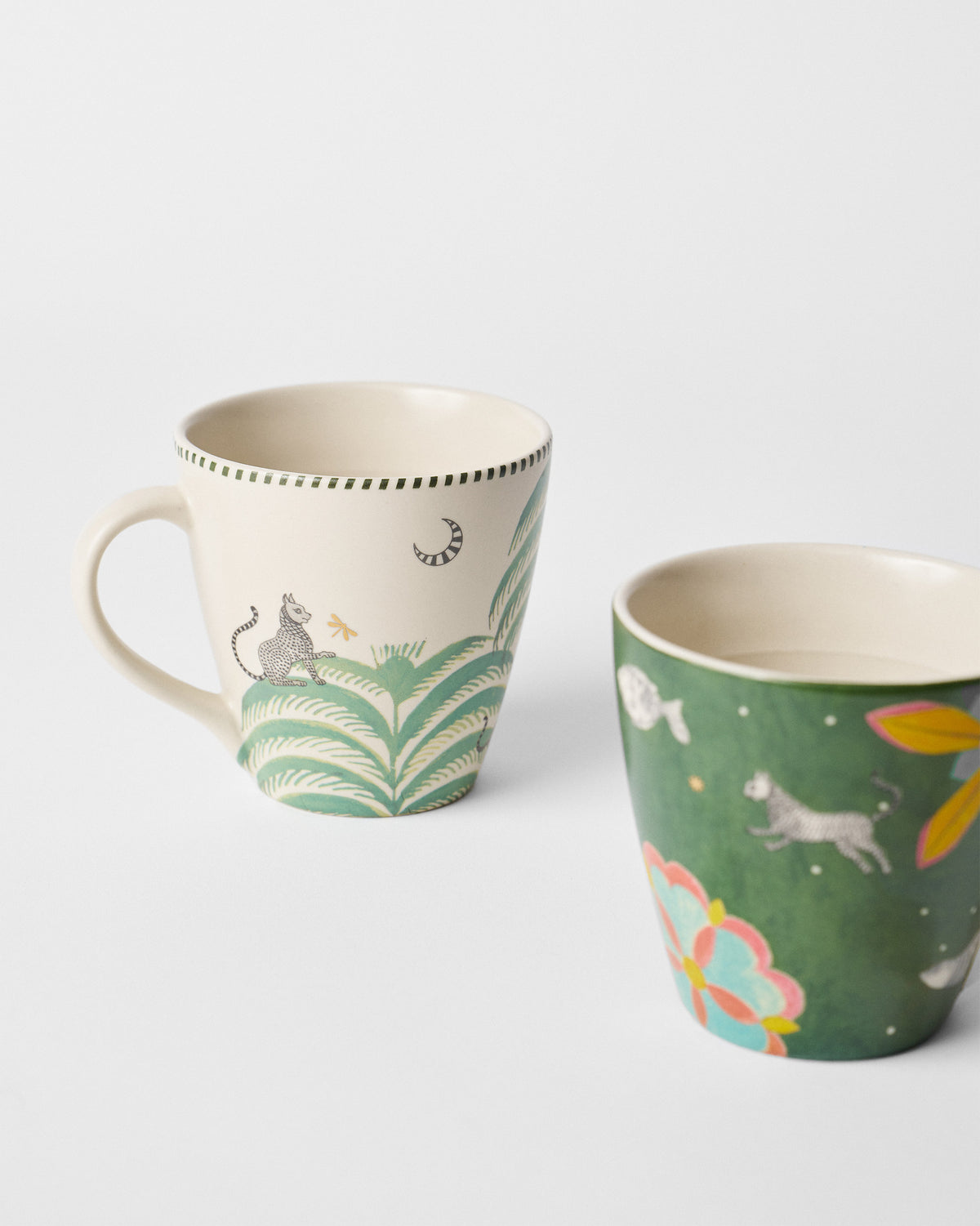 Cream and green Indus mugs showing a spotted cat, palms, and floral motifs.