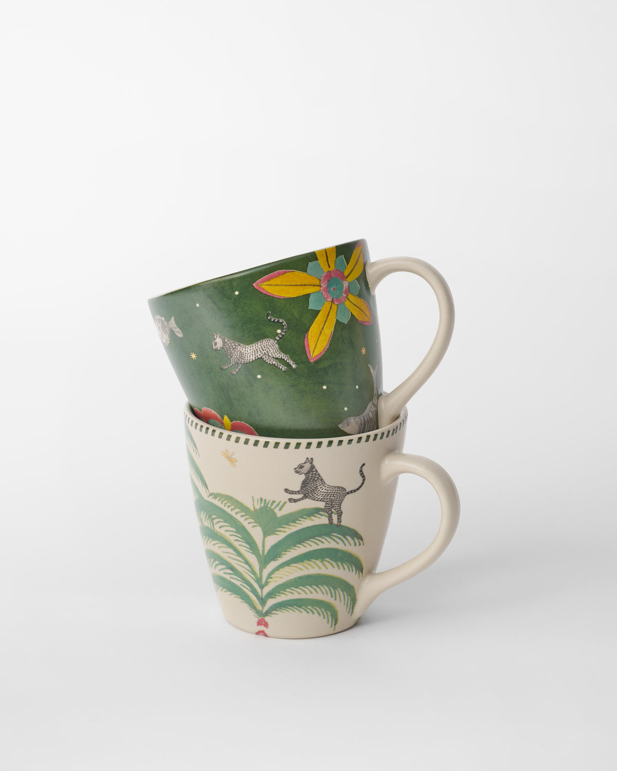 Stacked cream and green Indus mugs with spotted cats and tropical foliage.