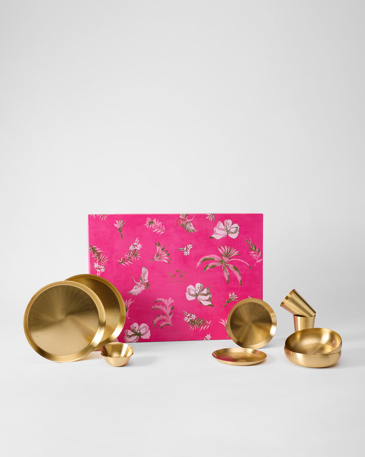 Madhu Dinner Gift Set of gold plates, bowls and cups beside a pink floral box.