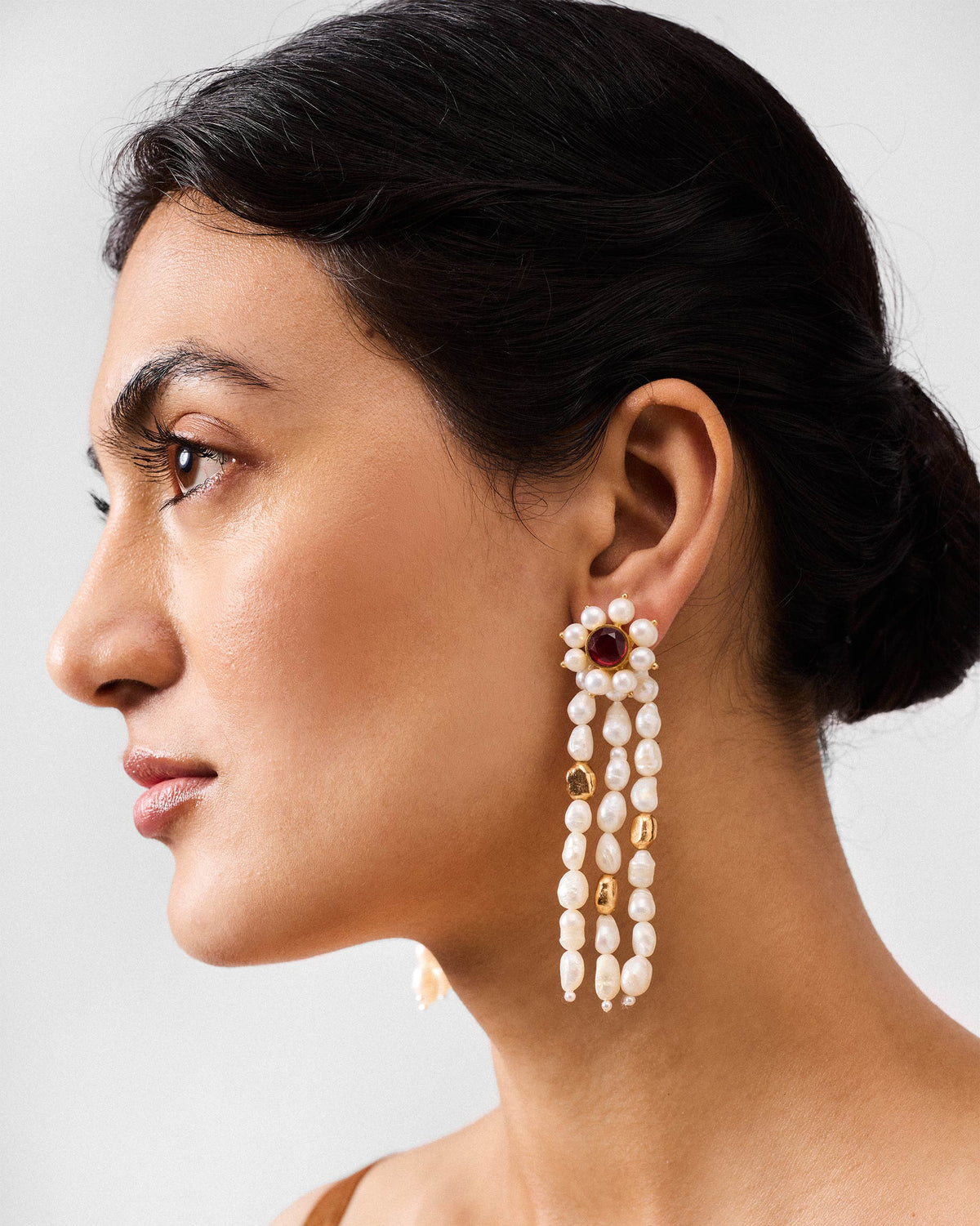Side profile of gold Meher earrings with pearl strands and a red stone on a person.