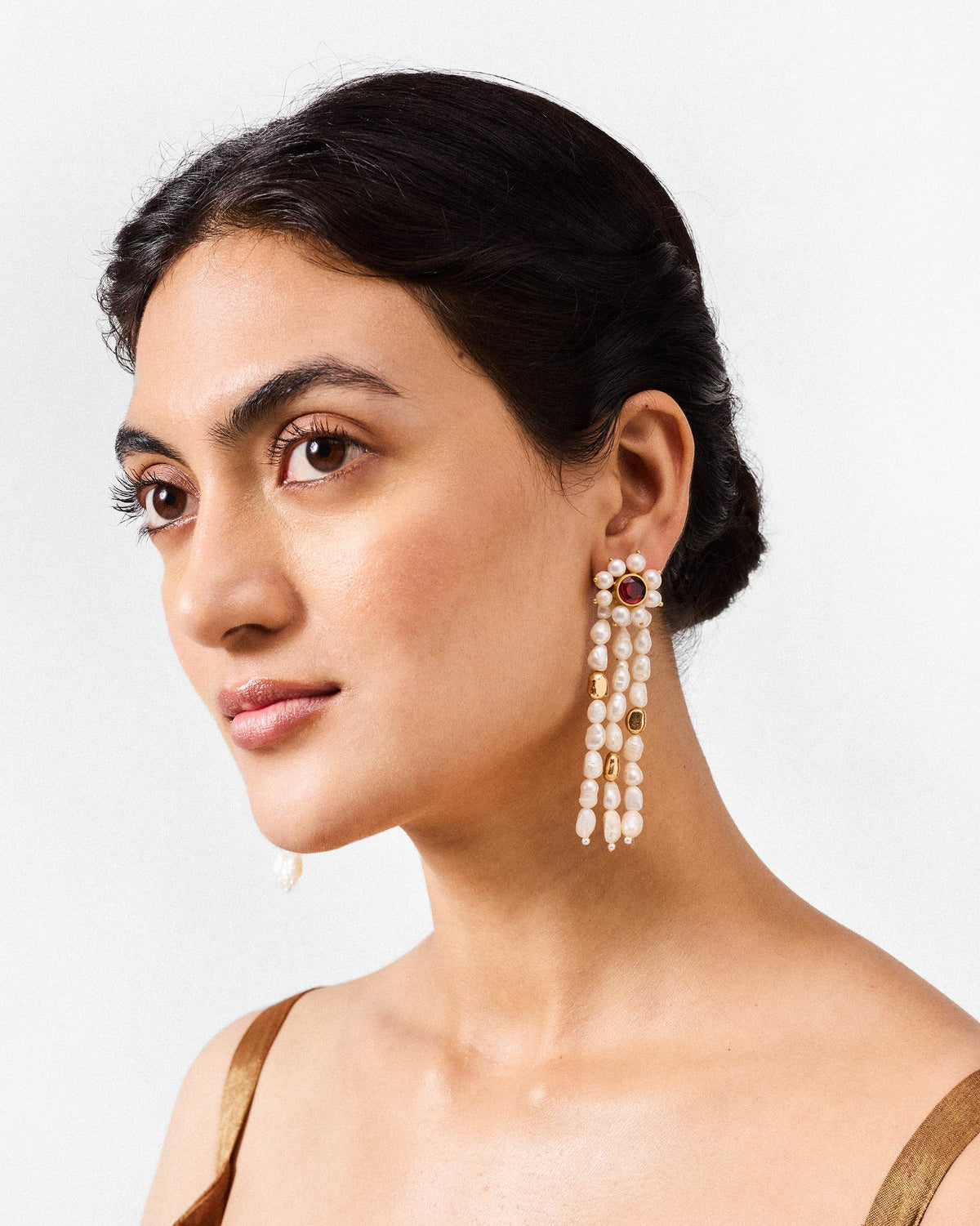 Person wearing gold Meher earrings with pearl strands and a red stone stud.