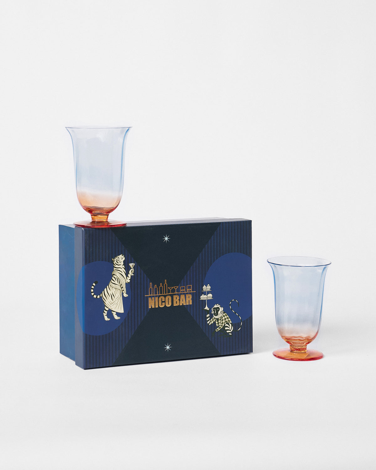 Oro cocktail glasses with blue-to-amber ombre beside a navy Nicobar gift box.
