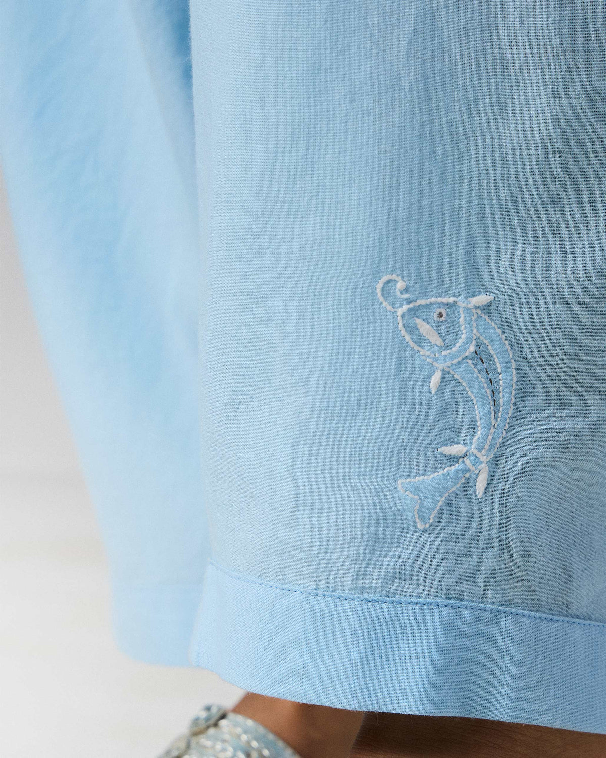 Close-up of a white embroidered fish near the hem of ice blue Aaira Pants.