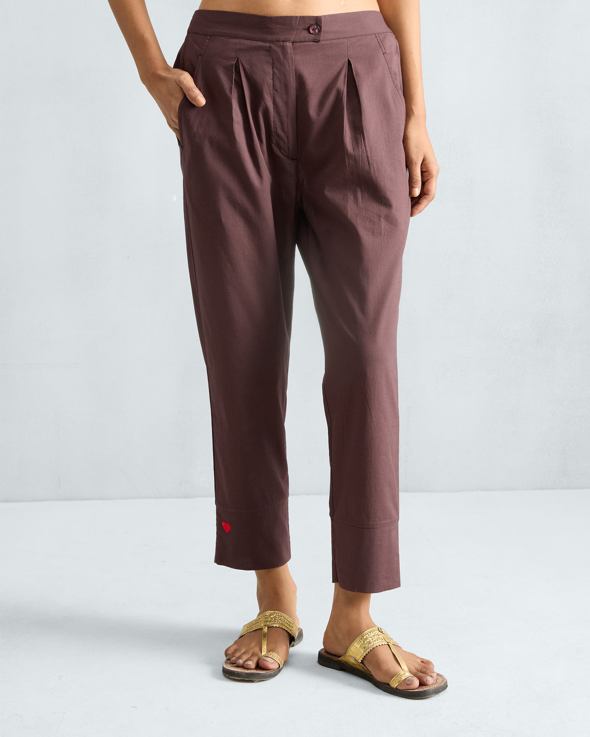 Person wearing chocolate plum narrow pants with front pleats and a red heart near the hem.