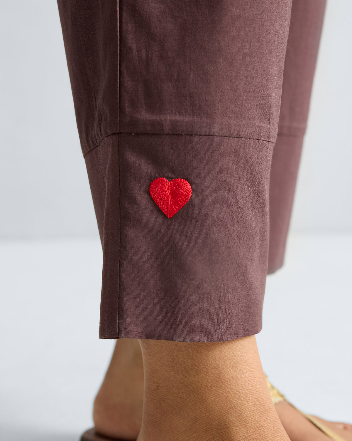 Close-up of a red embroidered heart on the hem of chocolate plum narrow pants.