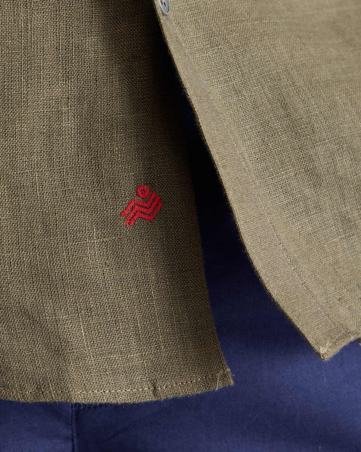 Close-up of red embroidery above the side vent of an olive linen shirt.