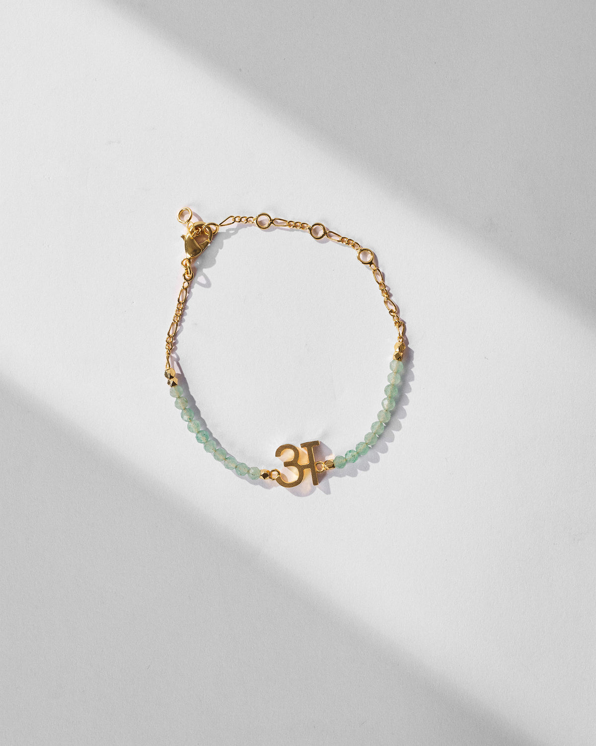 Akshar Bracelet (A) - Gold laid flat, with green beads and a gold charm.