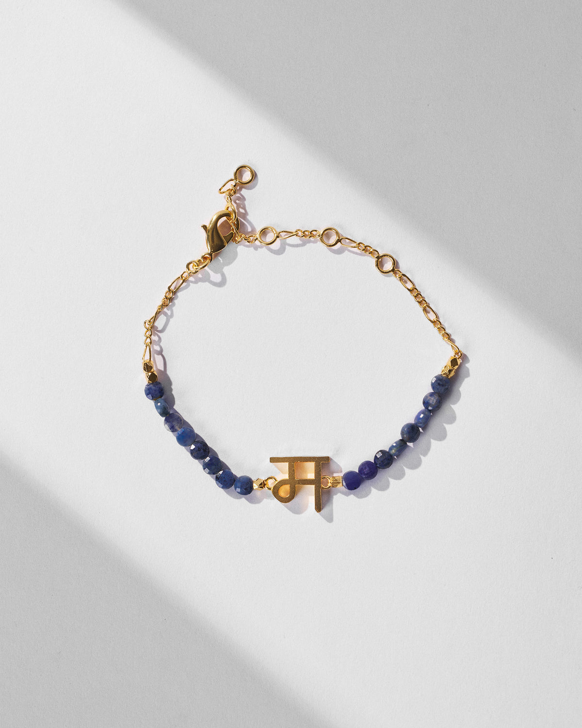 Akshar Bracelet (M) - Gold with blue beads and a gold charm, laid flat on a white surface.