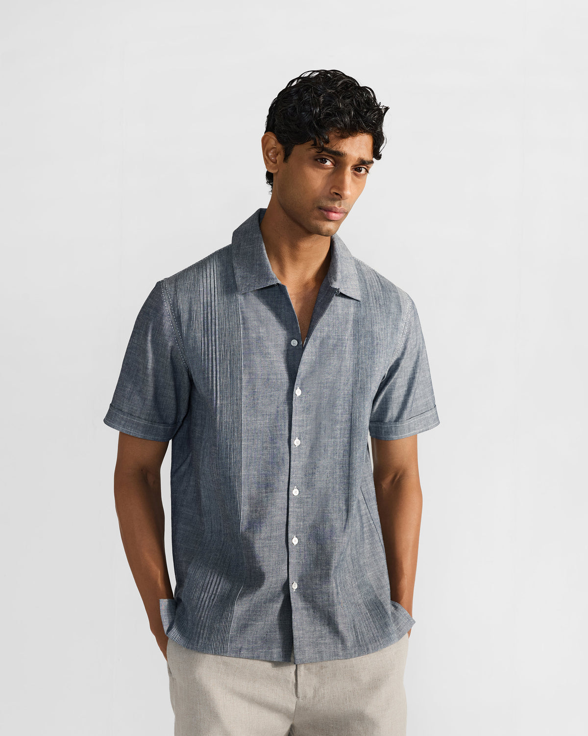 Man in an indigo textured short-sleeve shirt with vertical front pintucks and a camp collar, worn with light trousers.