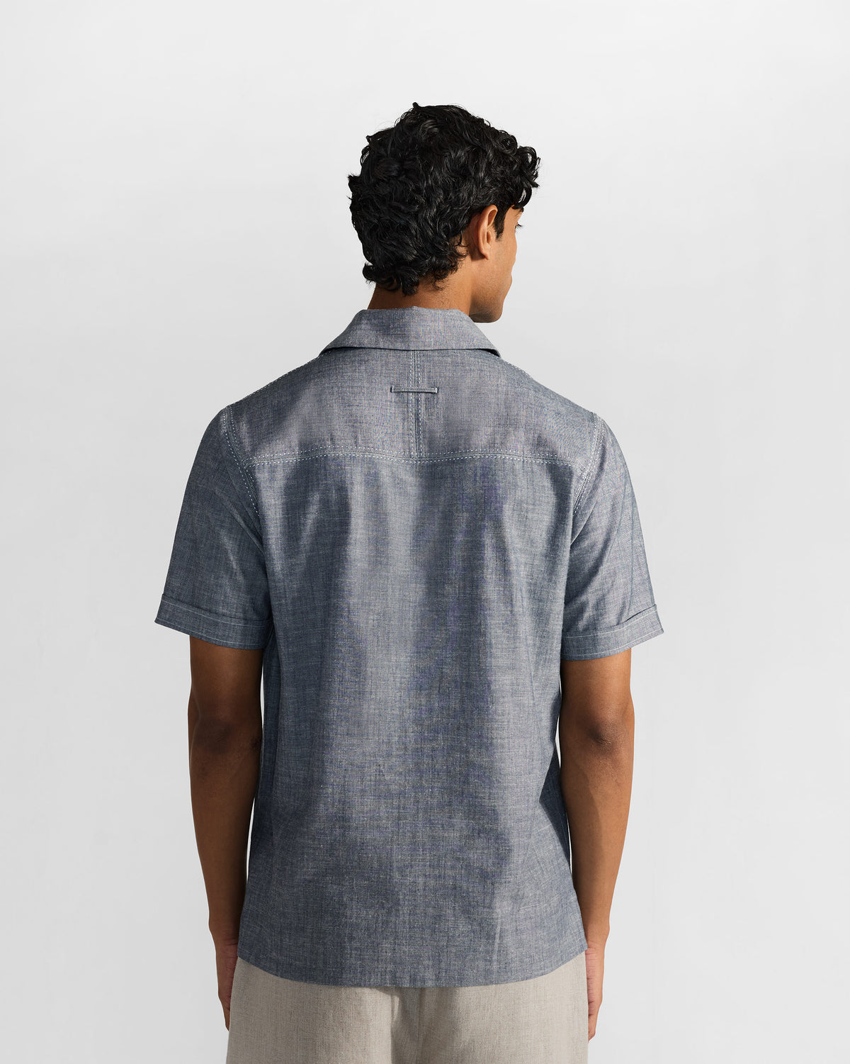 Back view of a man in an indigo short-sleeve shirt with a clean shoulder yoke and light trousers.