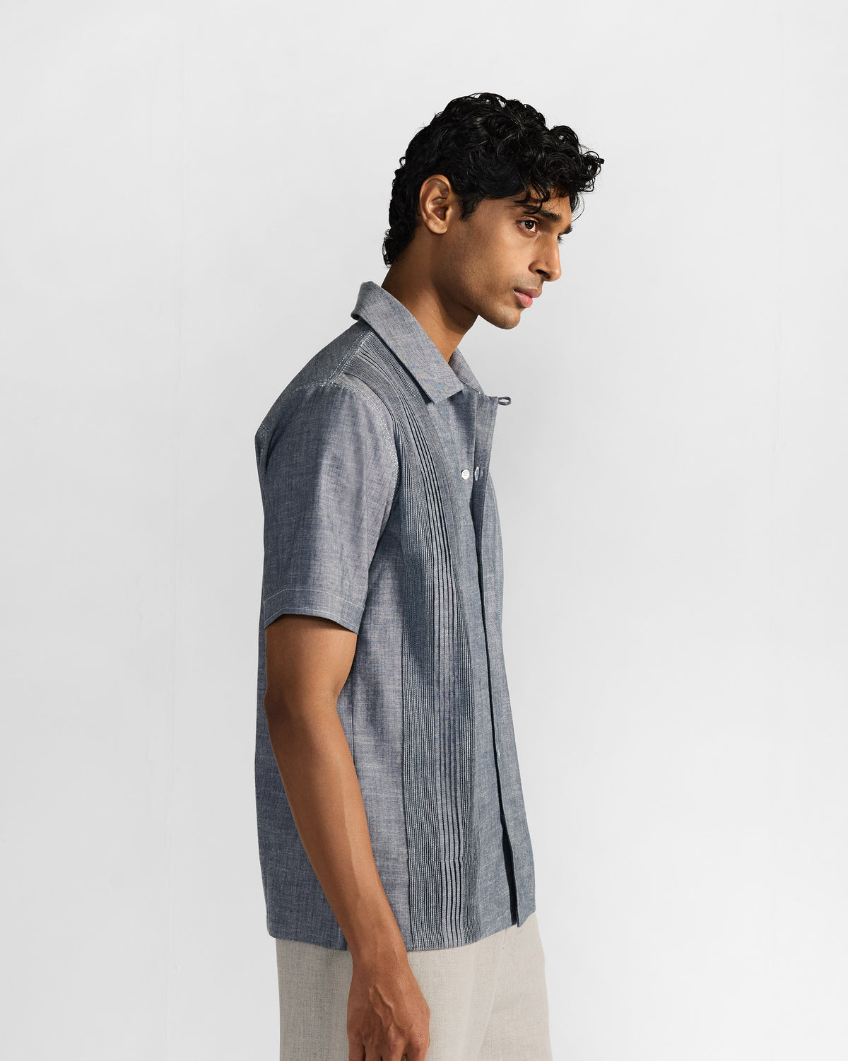 Side view of a man in an indigo short-sleeve shirt with vertical front pintucks and light trousers.