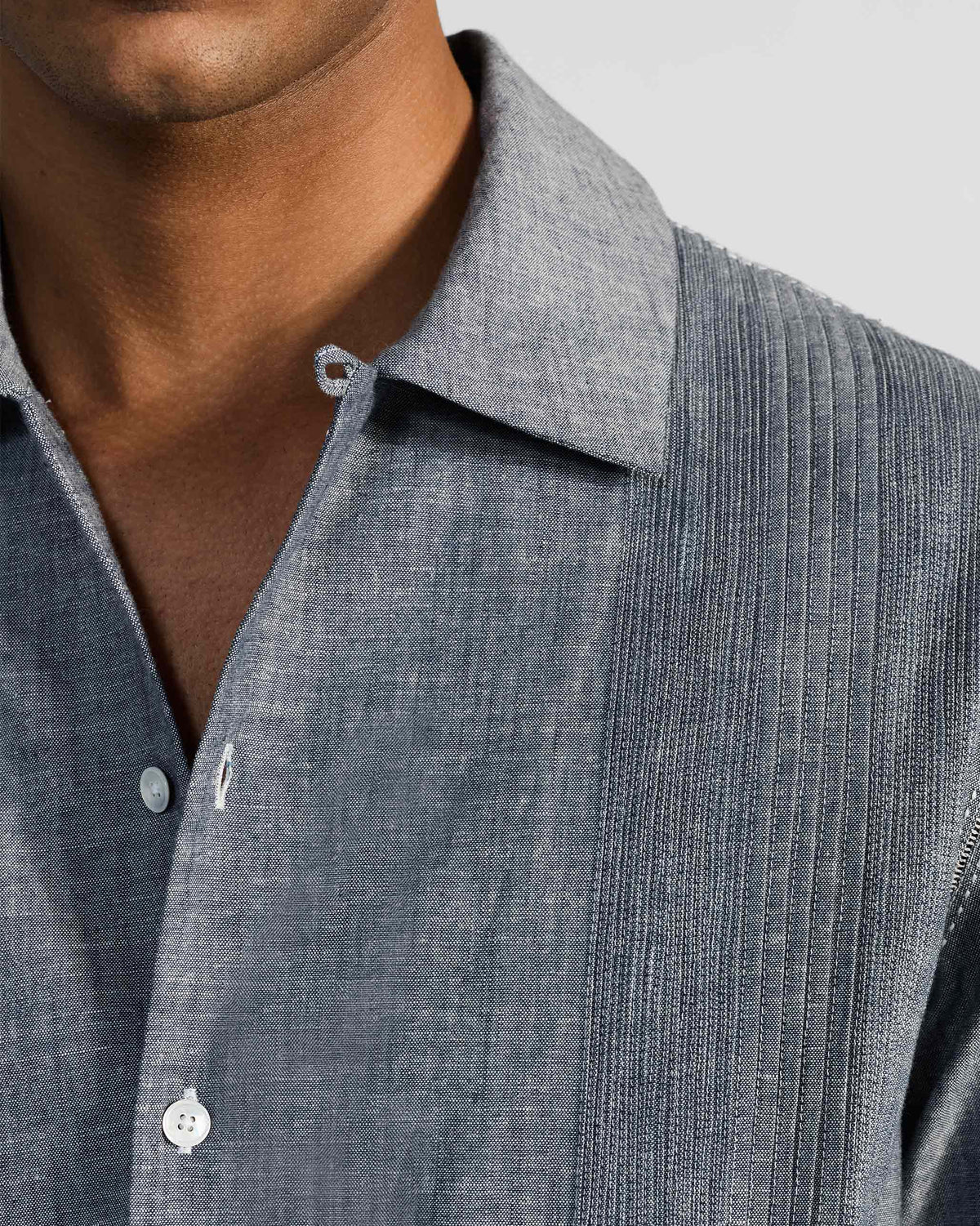 Close-up of an indigo textured shirt collar and white buttons beside a vertical pintuck panel.
