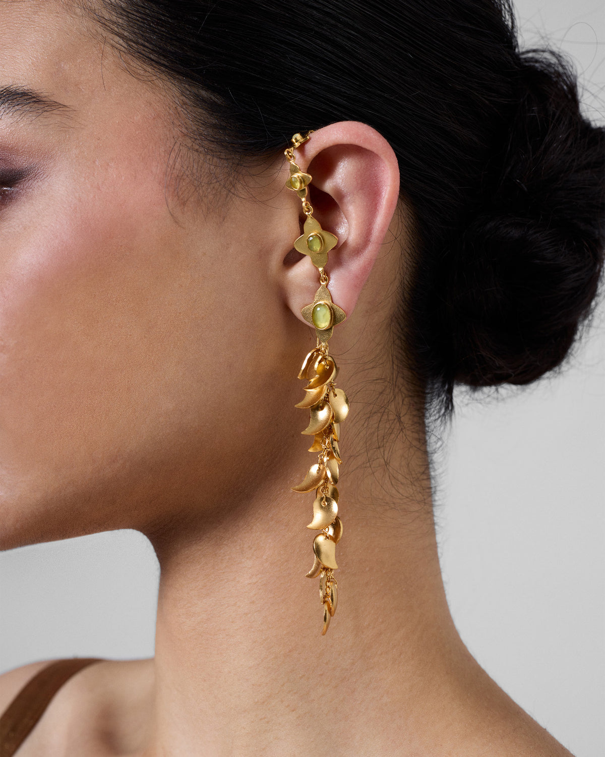 Person wearing a Kairi Earring with curved gold drops and green stones.