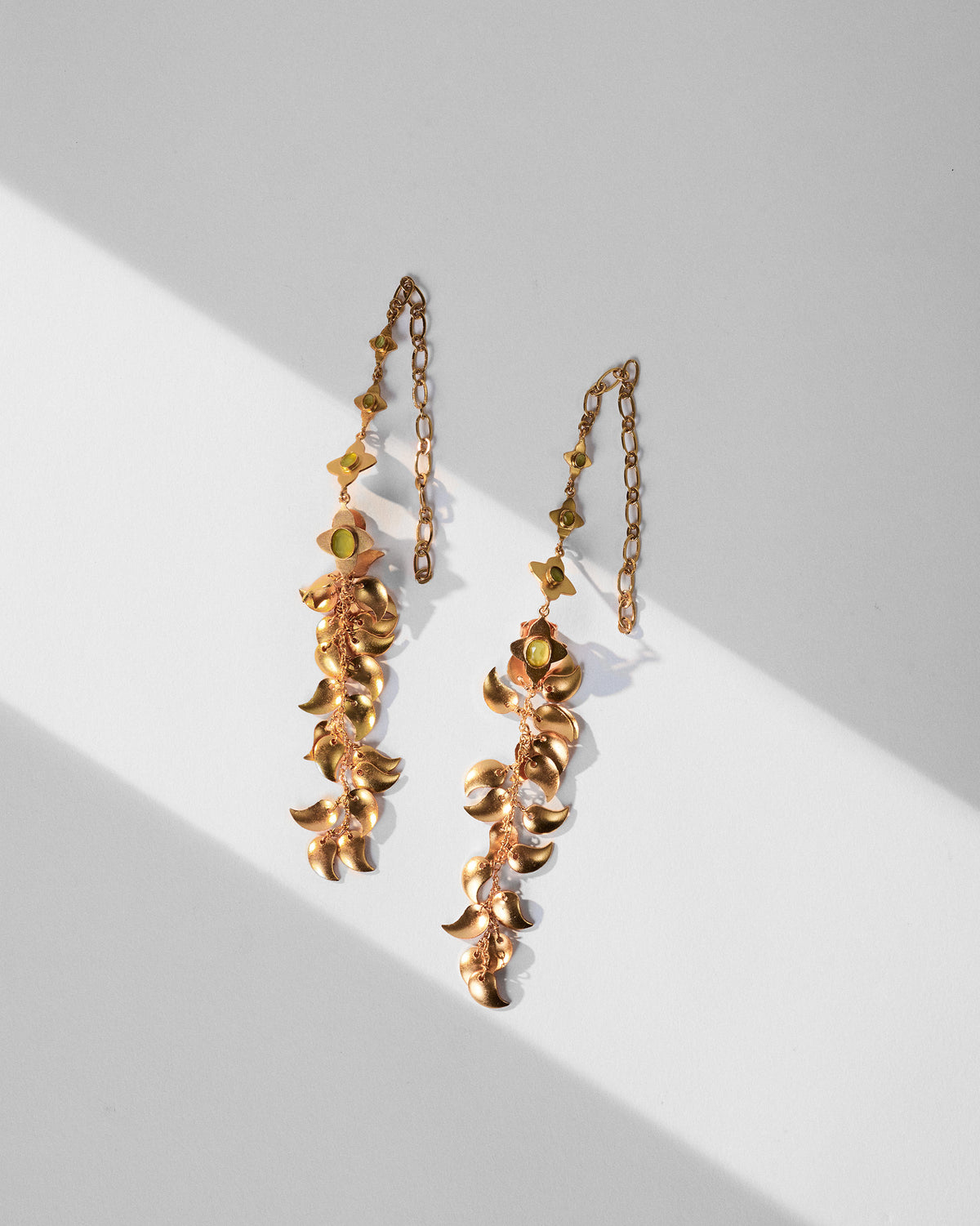 Pair of Kairi Earrings with curved gold drops and green stones, laid flat.