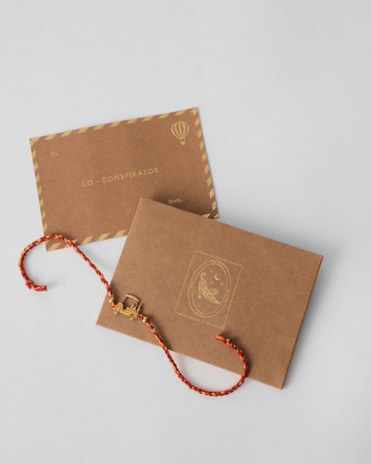 Golf Cart Rakhi in red and orange braid beside brown kraft envelopes.