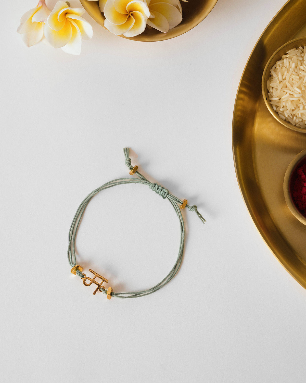 Alphabet M Rakhi with a sage green cord and gold charm, laid flat.