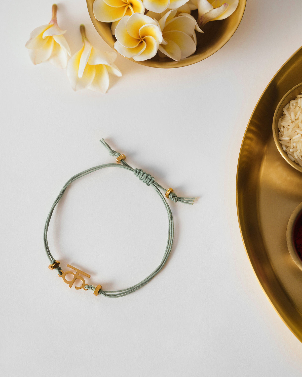 Alphabet K Rakhi with a sage green cord and gold charm, laid flat.