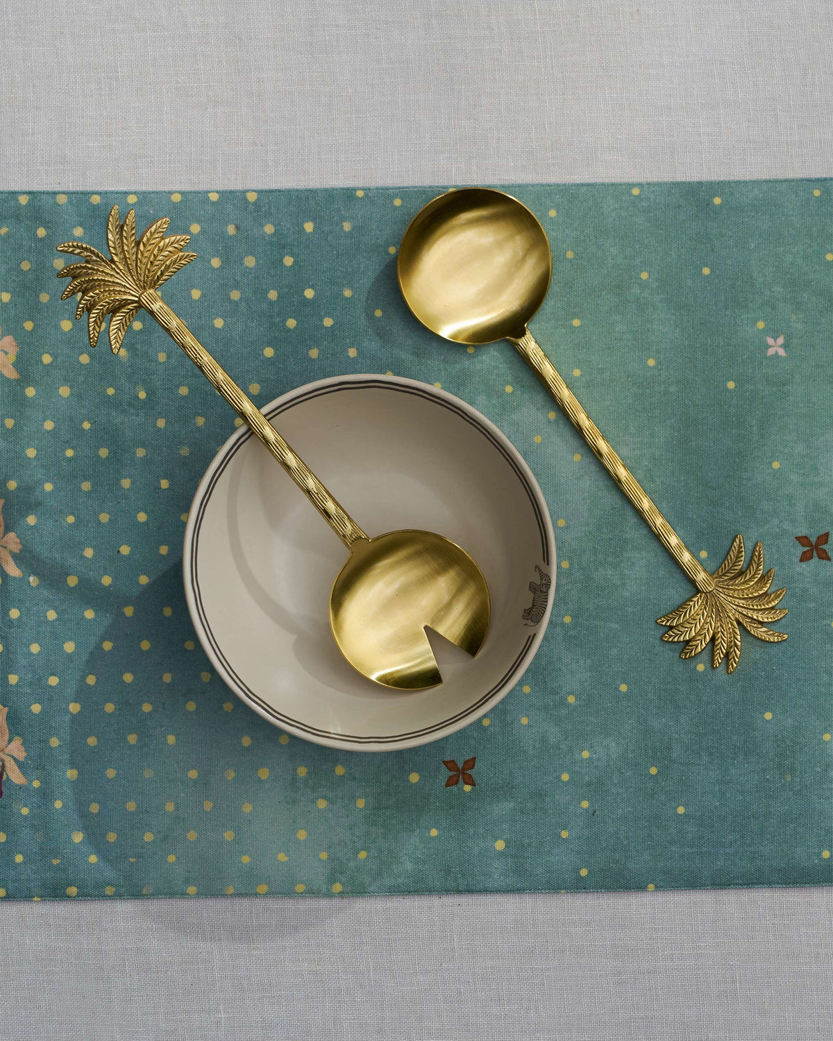 Gold Palm Bloom salad servers on a teal runner beside a small bowl.